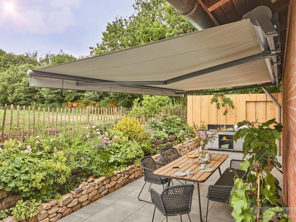 Gelenkarmmarkise mit beigem Tuch über sonniger Terrasse mit Essplatz und Garten