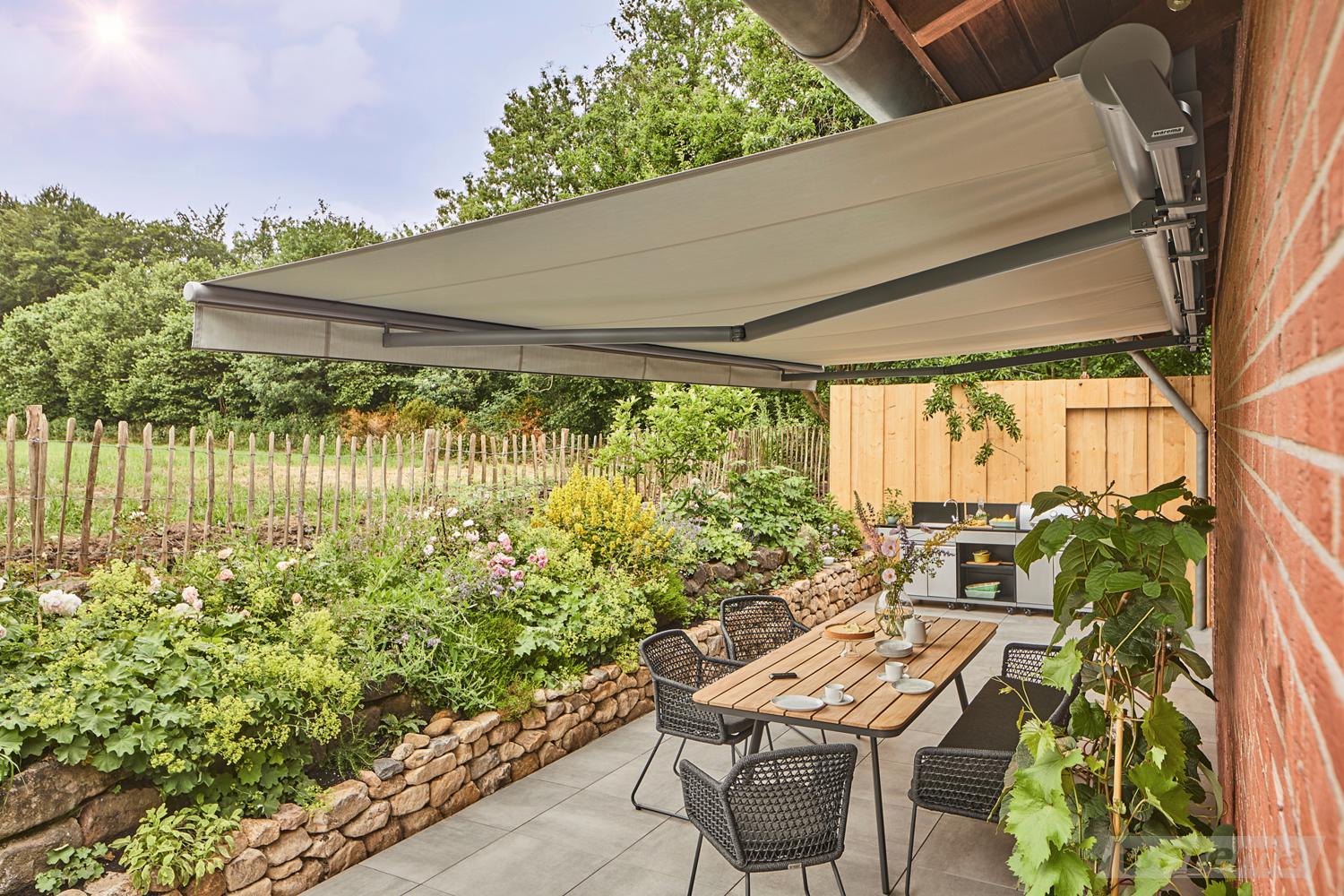 Gelenkarmmarkise mit beigem Tuch über sonniger Terrasse mit Essplatz und Garten