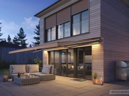 Moderne Gelenkarmmarkise an Holzfassade über Terrasse in der Abenddämmerung